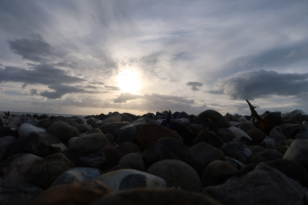 A view onto stones on a beach, in the background is water and a cloudy sky